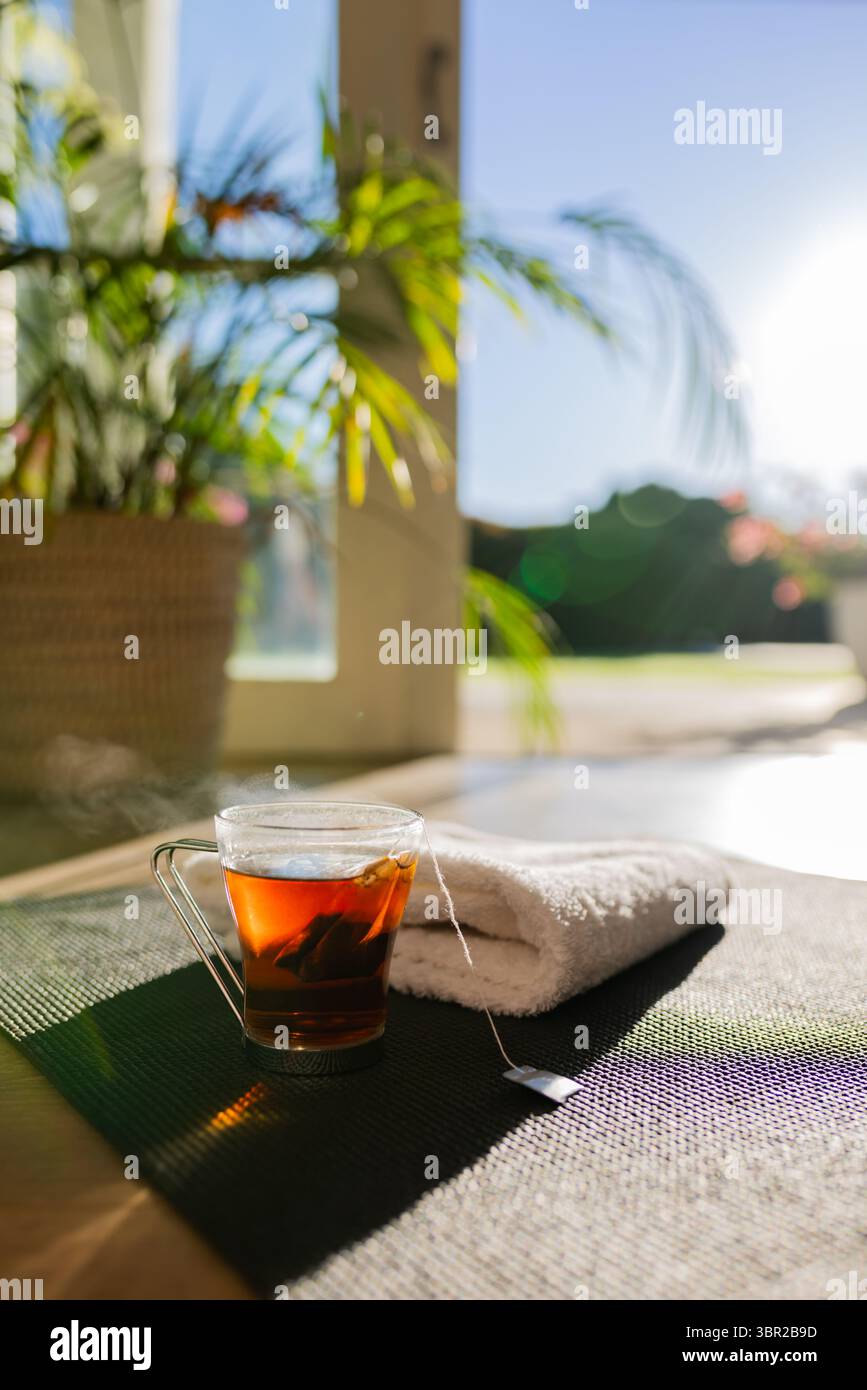 Tasse en verre thé ambré vapeur assis sur un tapis d'exercice sombre à côté d'une serviette roulée près de la porte coulissante. Bien-être, détente, sérénité, naturel, minimaliste, Banque D'Images