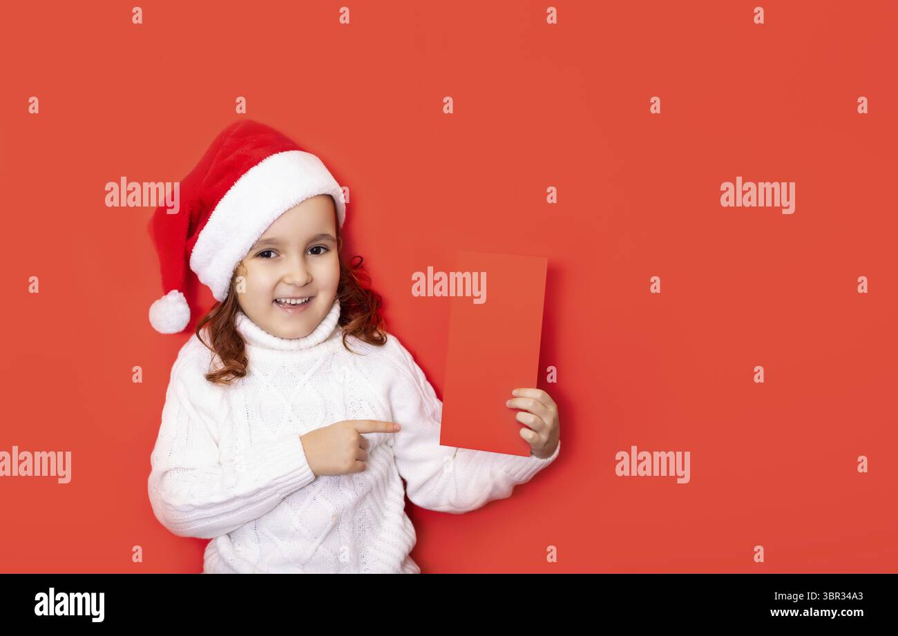 Nettes kaukasisches Maedchen mit roter Weihnachtsmannmuetze zeigt mit dem finger auf ein Zertifikat oder eine Grusskarte mit leerem Platz fuer Design Banque D'Images