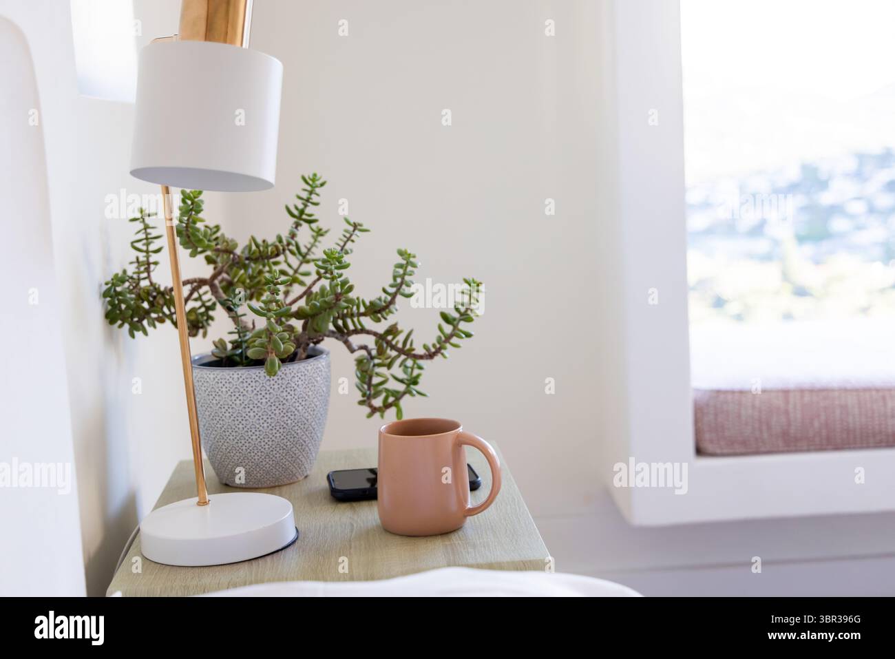 Lampe blanche avec tige dorée illumine la table de nuit avec succulente, tasse, téléphone et coussin de banc Banque D'Images