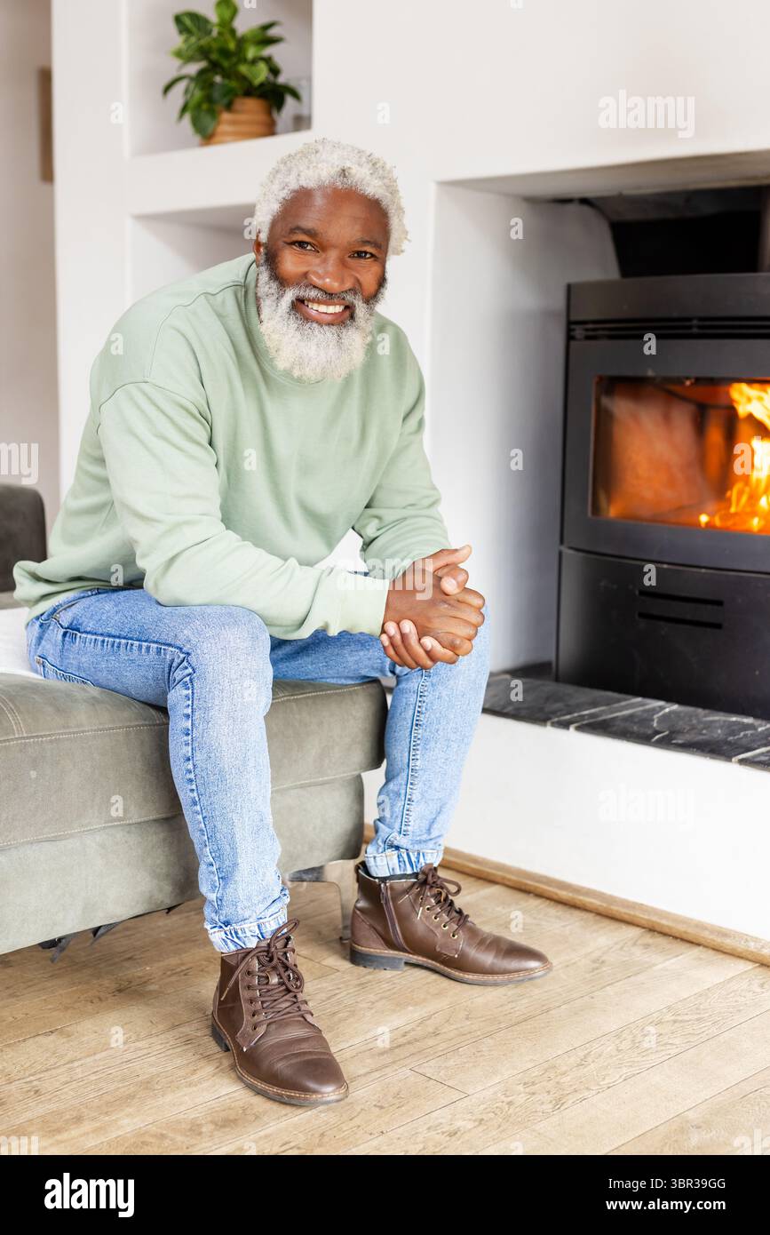 Homme afro-américain senior assis à la maison sur un canapé près d'une cheminée à bois avec des plantes en pot Banque D'Images