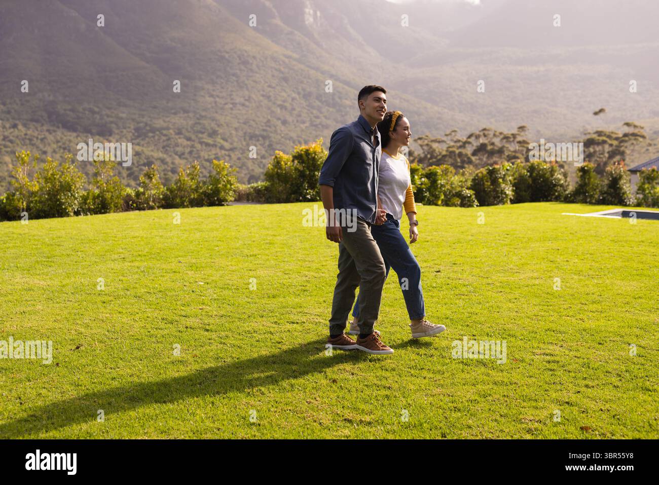Couple asiatique marchant côte à côte à travers la pelouse bien entretenue au parc avec des collines boisées en toile de fond Banque D'Images