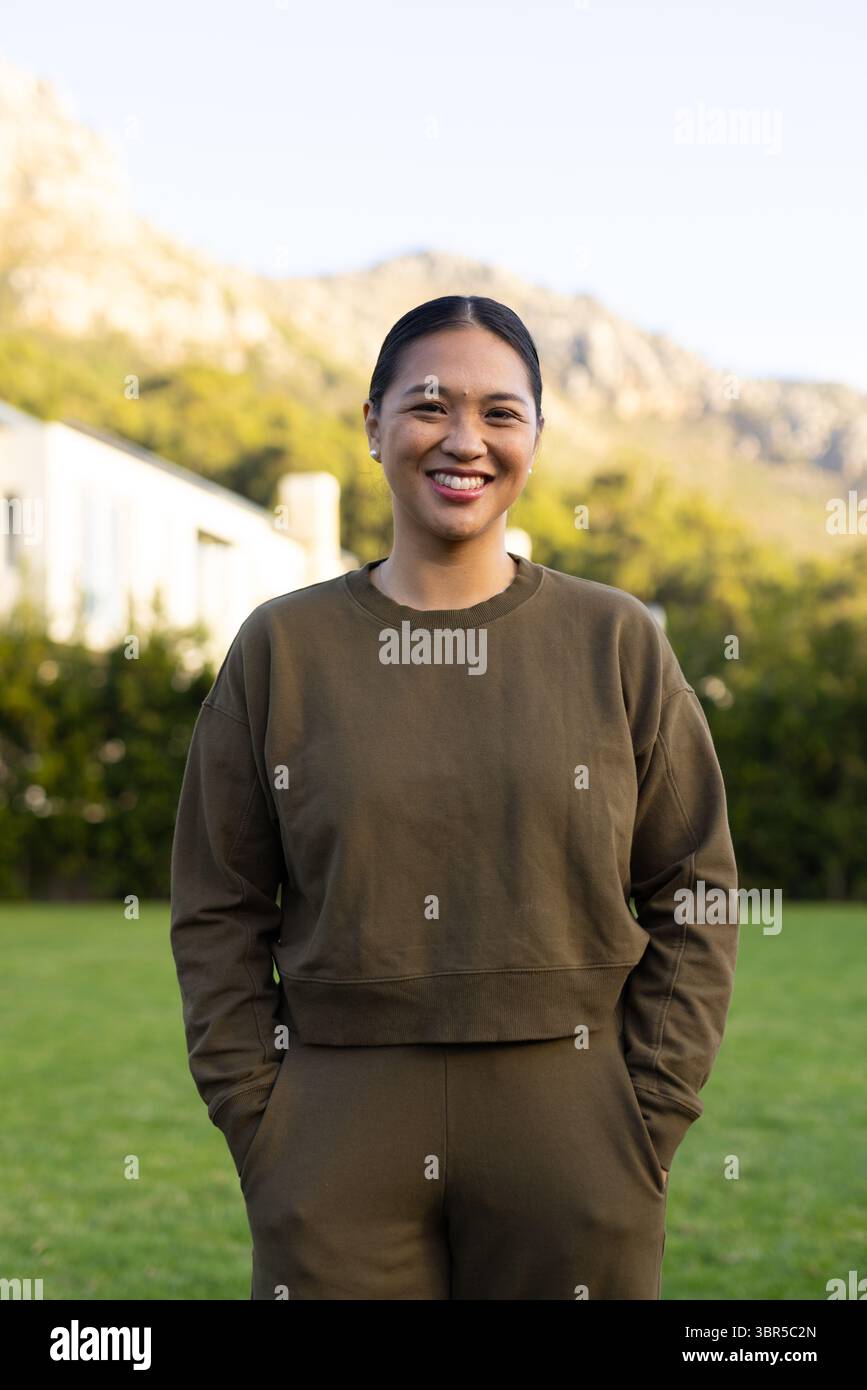 Femme asiatique souriante et debout sur la pelouse avant portant un survêtement vert olive près des haies de maison Banque D'Images