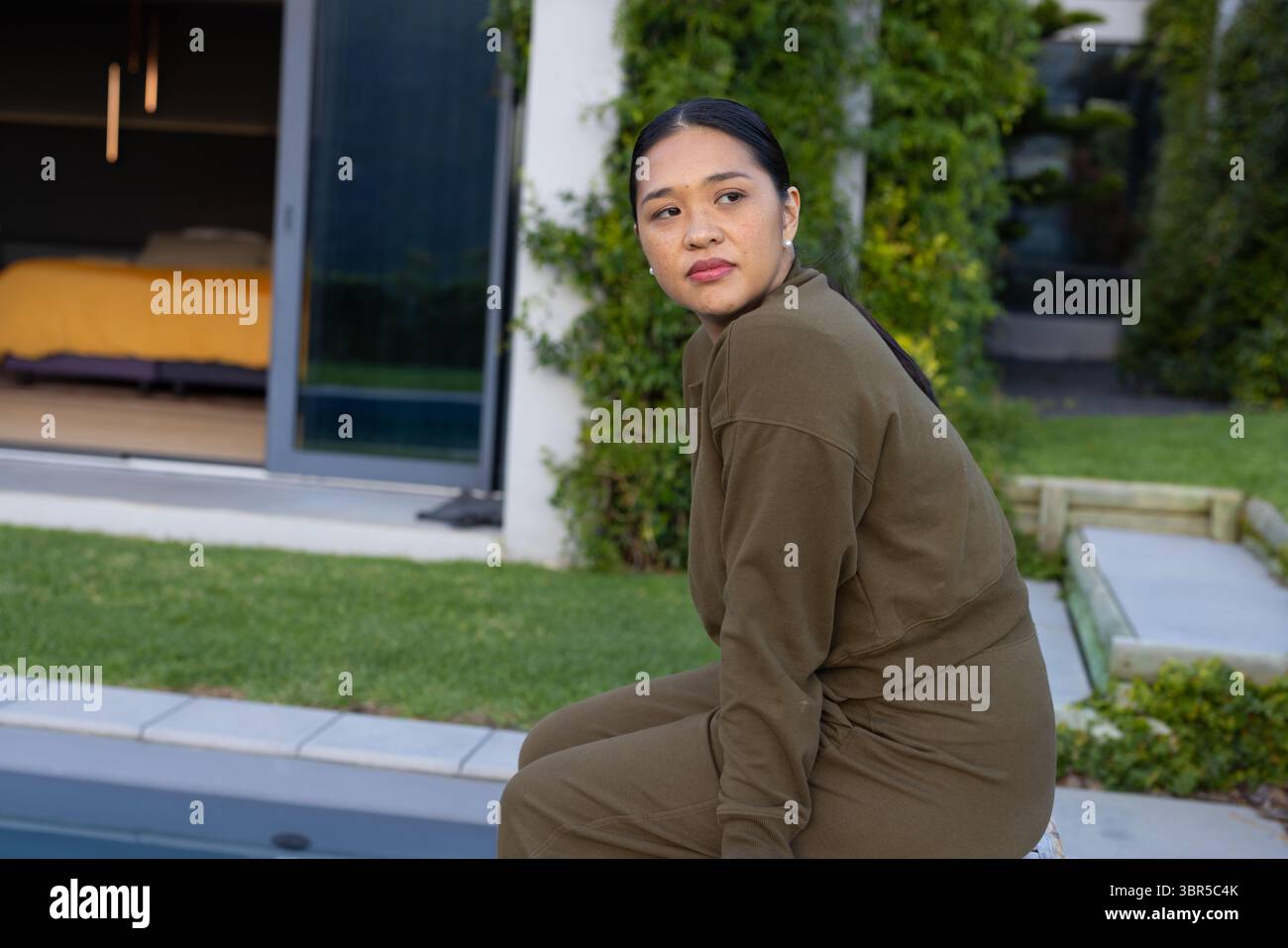 Femme asiatique assise sur le bord de la piscine en sweat-shirt vert olive sur la terrasse en pierre de cour arrière, espace de copie Banque D'Images