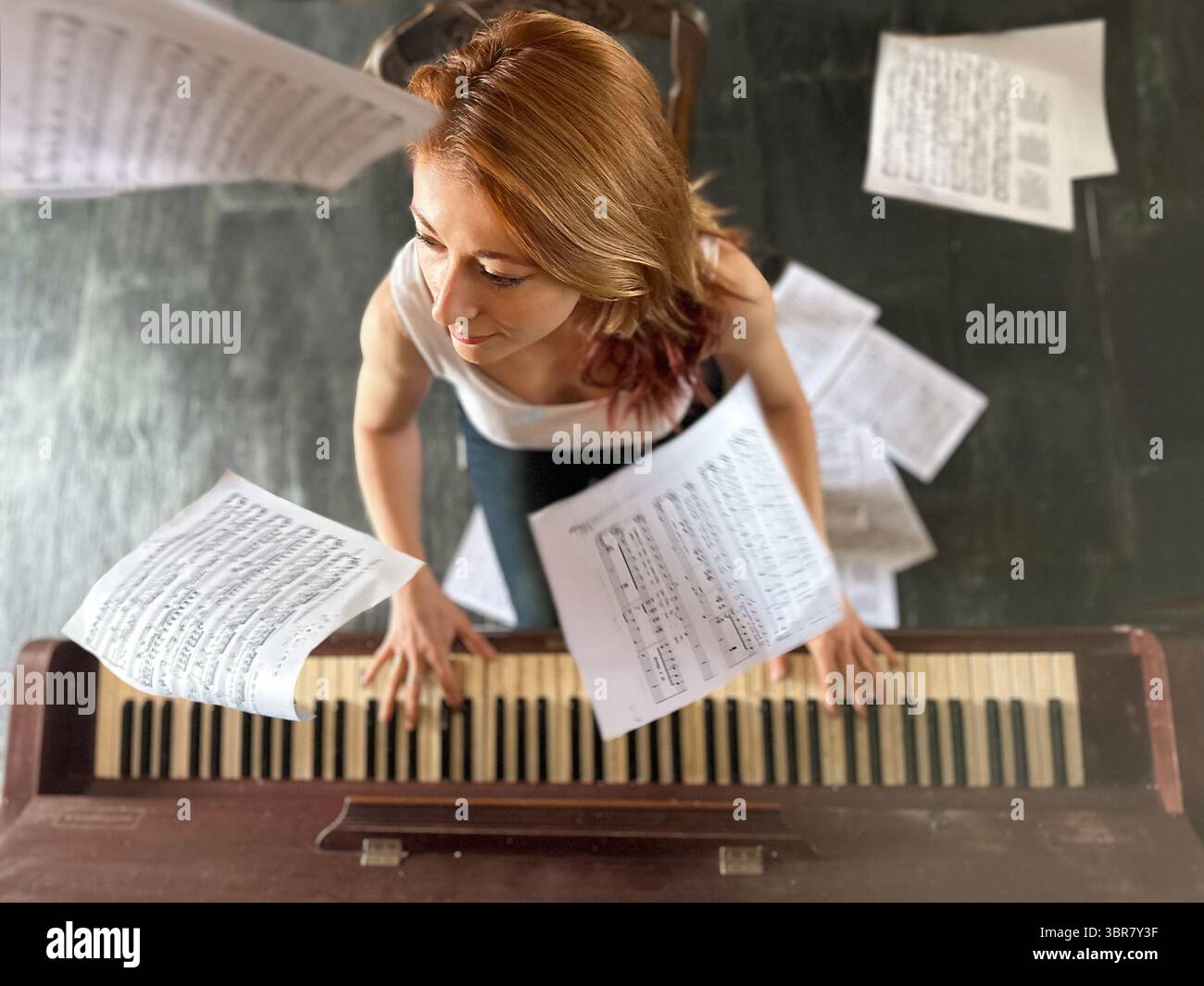 Femme aux cheveux de gingembre, 30 ans, jouant du piano à la maison. Vue en grand angle avec des partitions tombant autour d'elle et allongées sur le sol. Artistique, émotionnel, Banque D'Images