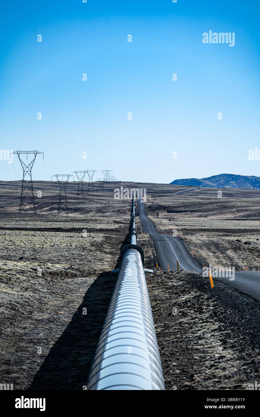 Vue d'un pipeline argenté s'étendant à travers le paysage aride, parallèle à une route sous un ciel bleu clair, Nesjavellir, Grímsnes- og Grafningshreppur, Islande. Banque D'Images