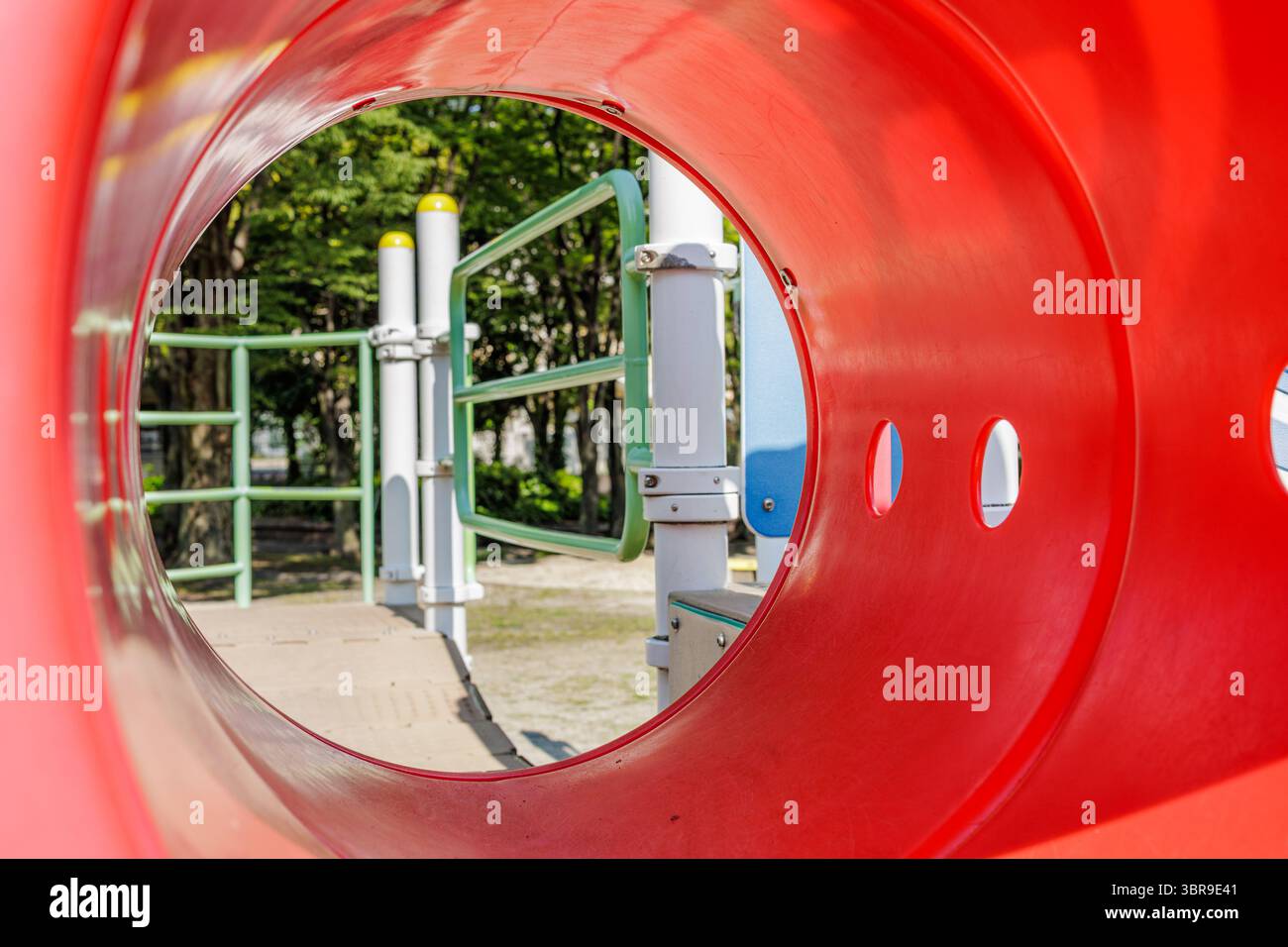 Vue rapprochée à travers le tunnel rouge vif d'un terrain de jeu, observant les structures environnantes et la verdure au-delà. La lumière du soleil ajoute des ombres douces. Banque D'Images