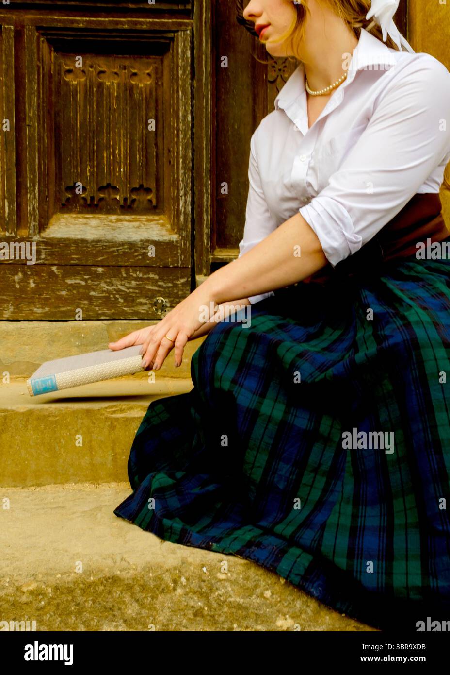 Une femme en chemise blanche, collier de perles et jupe à carreaux est assise sur les marches, tenant un livre. Banque D'Images