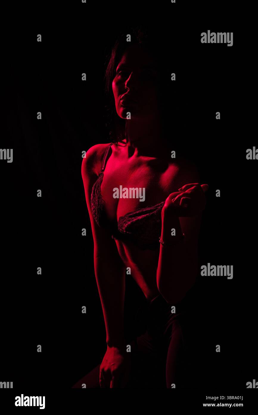 Mannequin prend une pose confiante dans un cadre sombre éclairé par une lumière rouge, soulignant sa silhouette et créant une atmosphère séduisante L'ambiance transmet un sentiment de mystère et d'élégance Banque D'Images