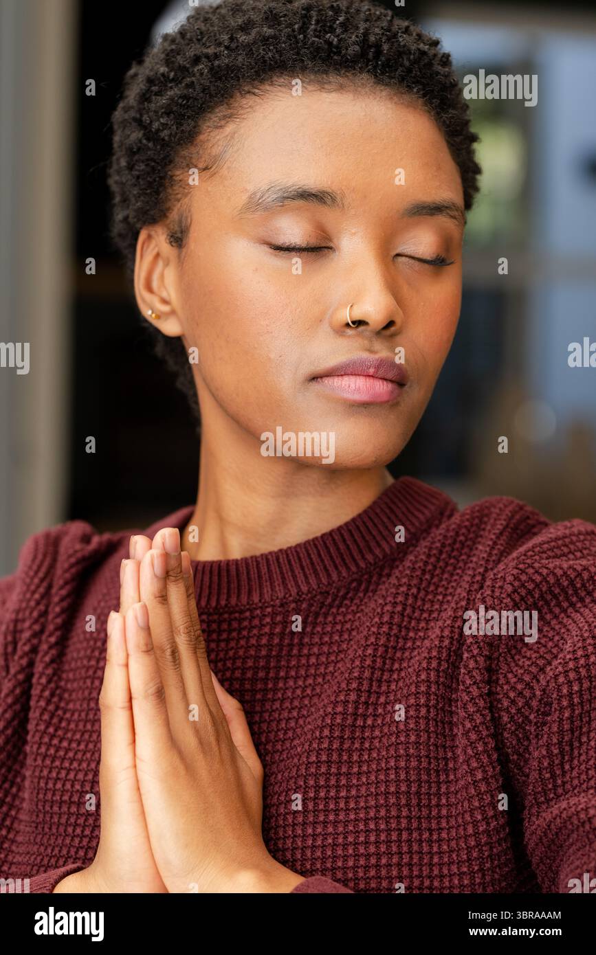 Femme afro-américaine méditant en pressant les mains à la maison portant un arceau de nez et des boucles d'oreilles d'arceau Banque D'Images