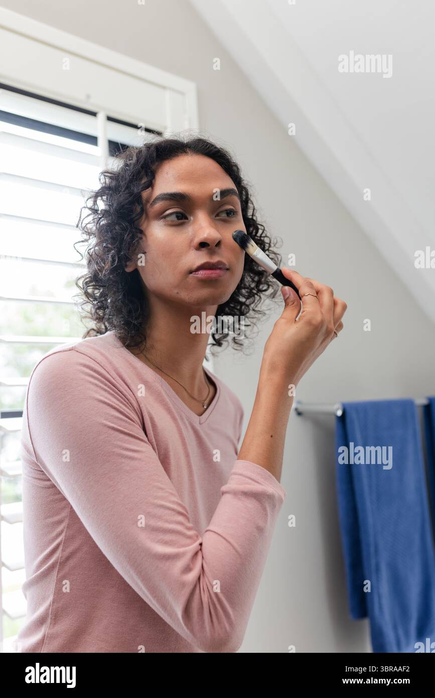 Personne non binaire appliquant un pinceau de maquillage à la joue dans la salle de bain près des volets blancs et serviette bleue Banque D'Images