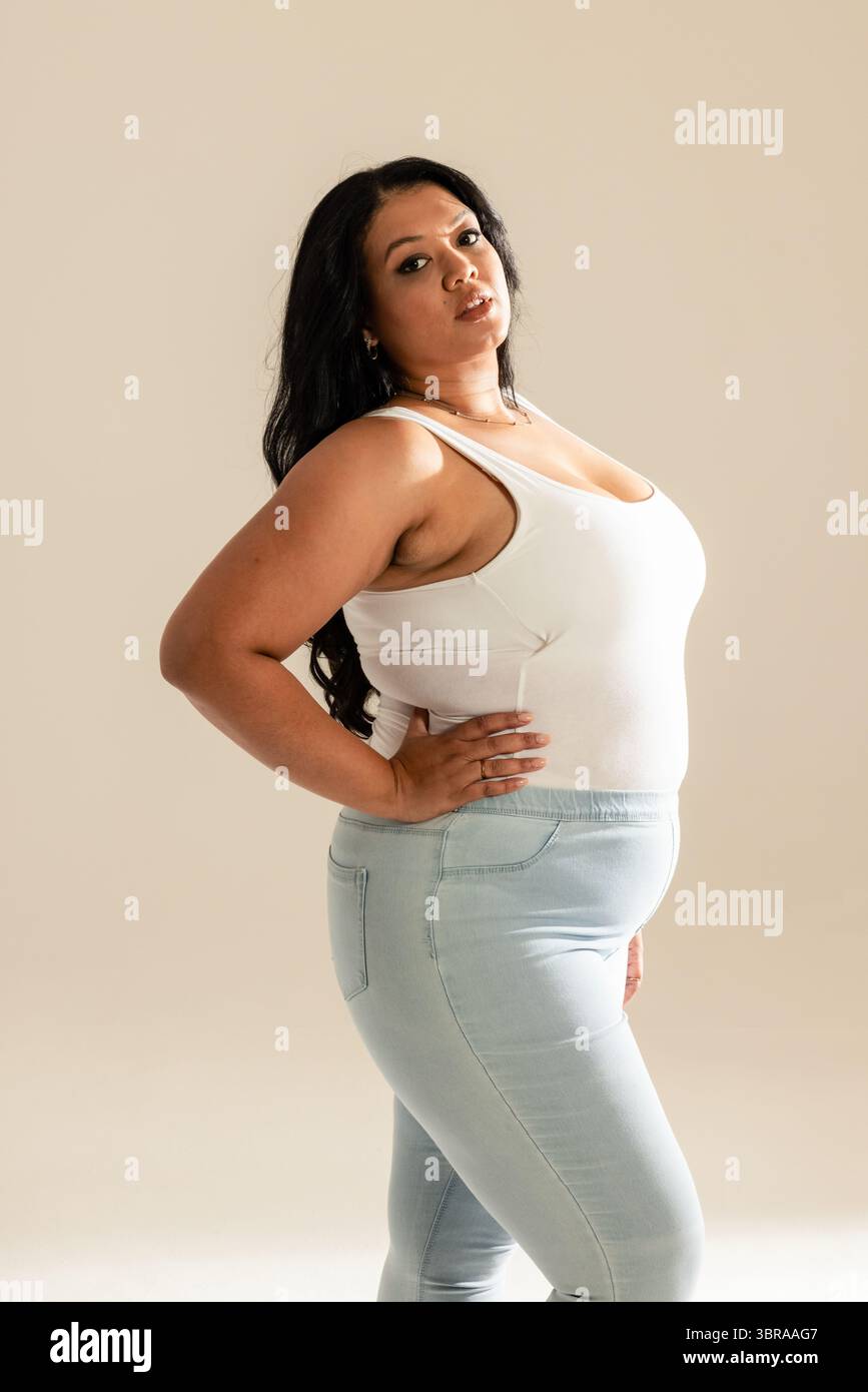 Femme posant sur fond beige en studio portant un débardeur blanc et un Jean avec collier et boucles d'oreilles Banque D'Images