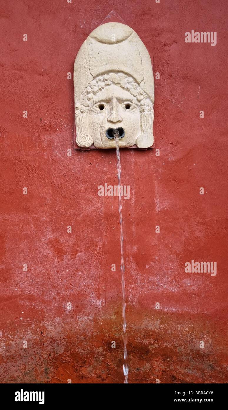 Une sculpture de masque de style classique haut sur un mur de plâtre rouge, l'eau coule d'un tuyau dans la bouche. Banque D'Images