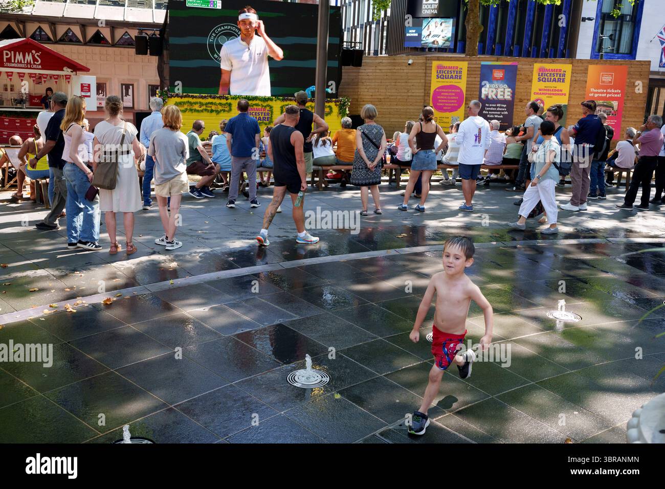 Alors que la canicule britannique de juillet se poursuit dans le week-end, avec des avertissements de chaleur de met Office Amber en place pour le sud-est, le public regarde le match de demi-finale de Wimbledon entre Taylor Fritz et Carlos Alcaraz tandis que les enfants éclaboussent dans les eaux rafraîchissantes des fontaines de Leicester Square, le 11 juillet 2025, à Londres, en Angleterre. Banque D'Images