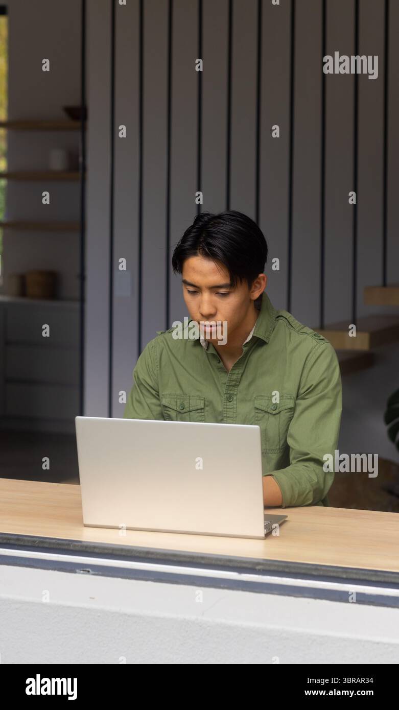 Homme asiatique portant chemise vert olive tapant sur ordinateur portable assis au comptoir en bois dans le bureau Banque D'Images
