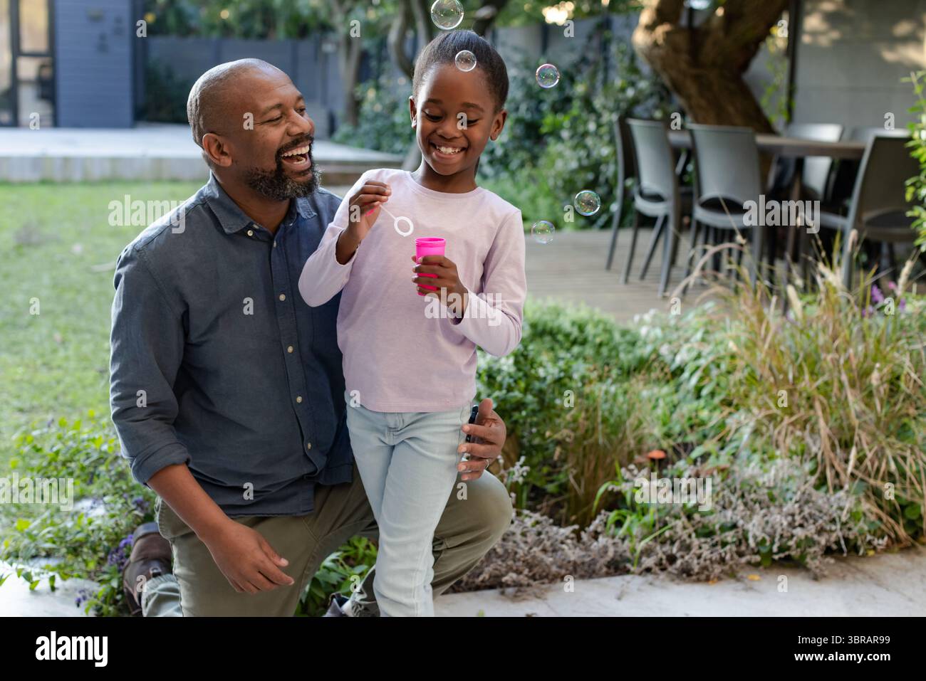 Père noir agenouillé à côté de sa fille soufflant des bulles du récipient rose sur le patio Banque D'Images