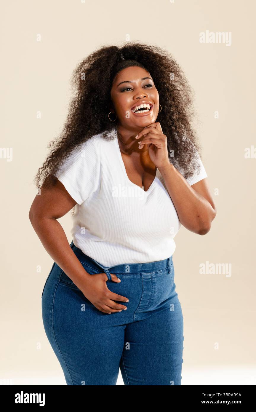 Femme afro-américaine souriante montrant des cheveux bouclés dans un studio de photographie portant un haut blanc et un Jean Banque D'Images