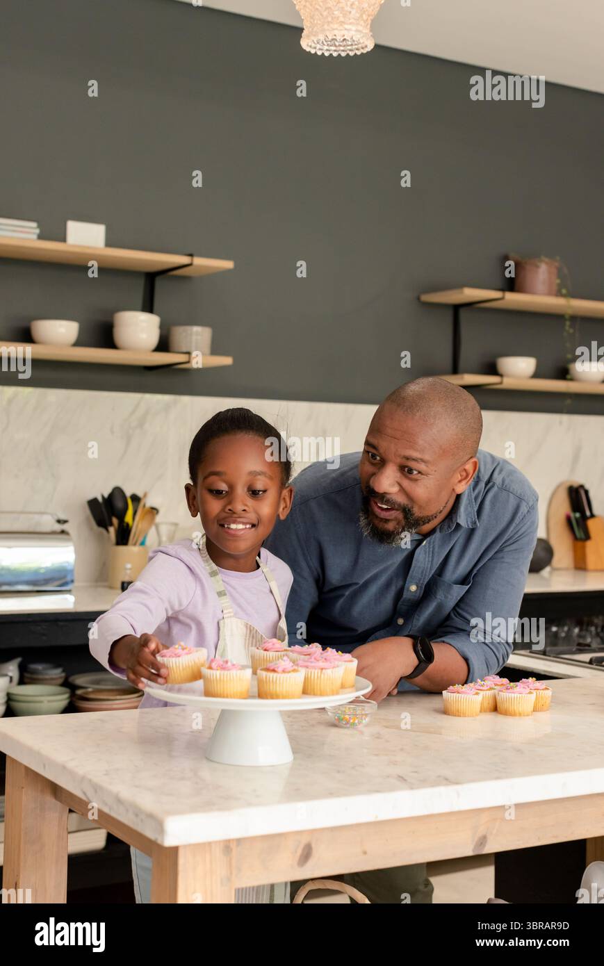 Papa afro-américain et sa fille décorant des cupcakes roses sur un stand à gâteau à l'îlot de cuisine en marbre Banque D'Images