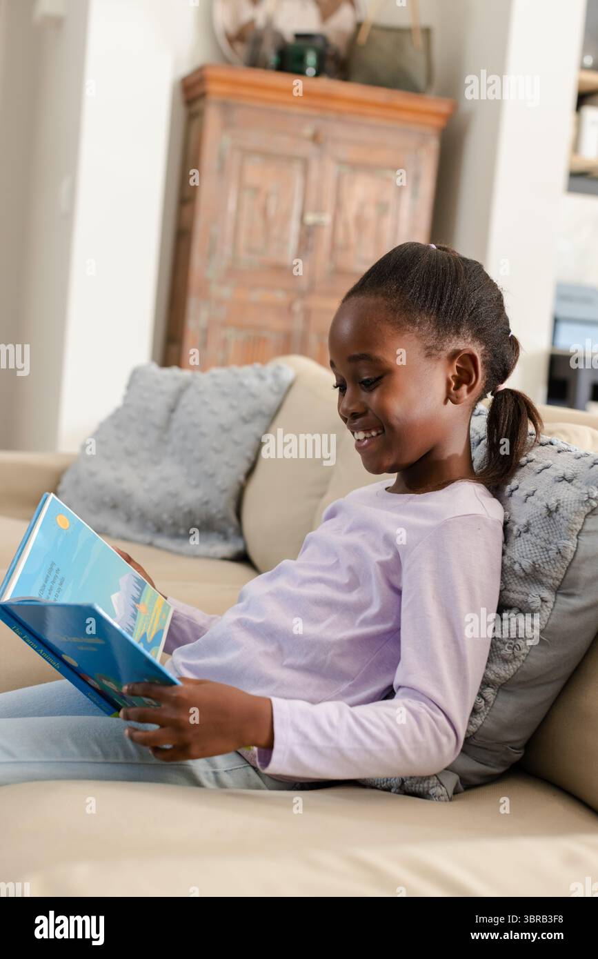 Enfant afro-américaine lisant un livre à couverture rigide bleu vibrant sur un canapé beige à la maison, espace de copie Banque D'Images