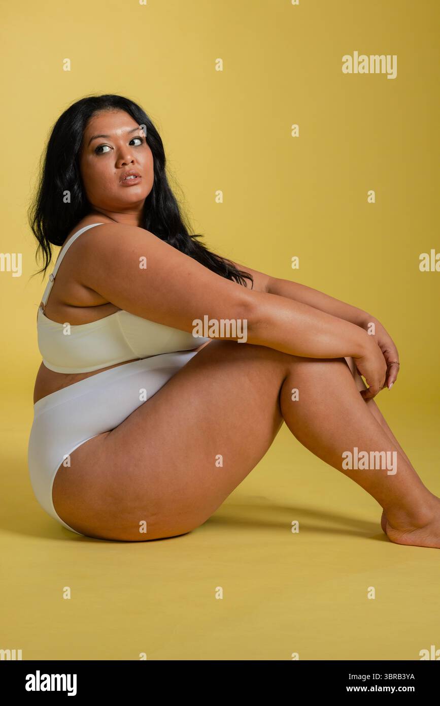 Femme assise serrant les genoux tout en portant un soutien-gorge blanc et un slip sur un sol de studio jaune minimaliste Banque D'Images