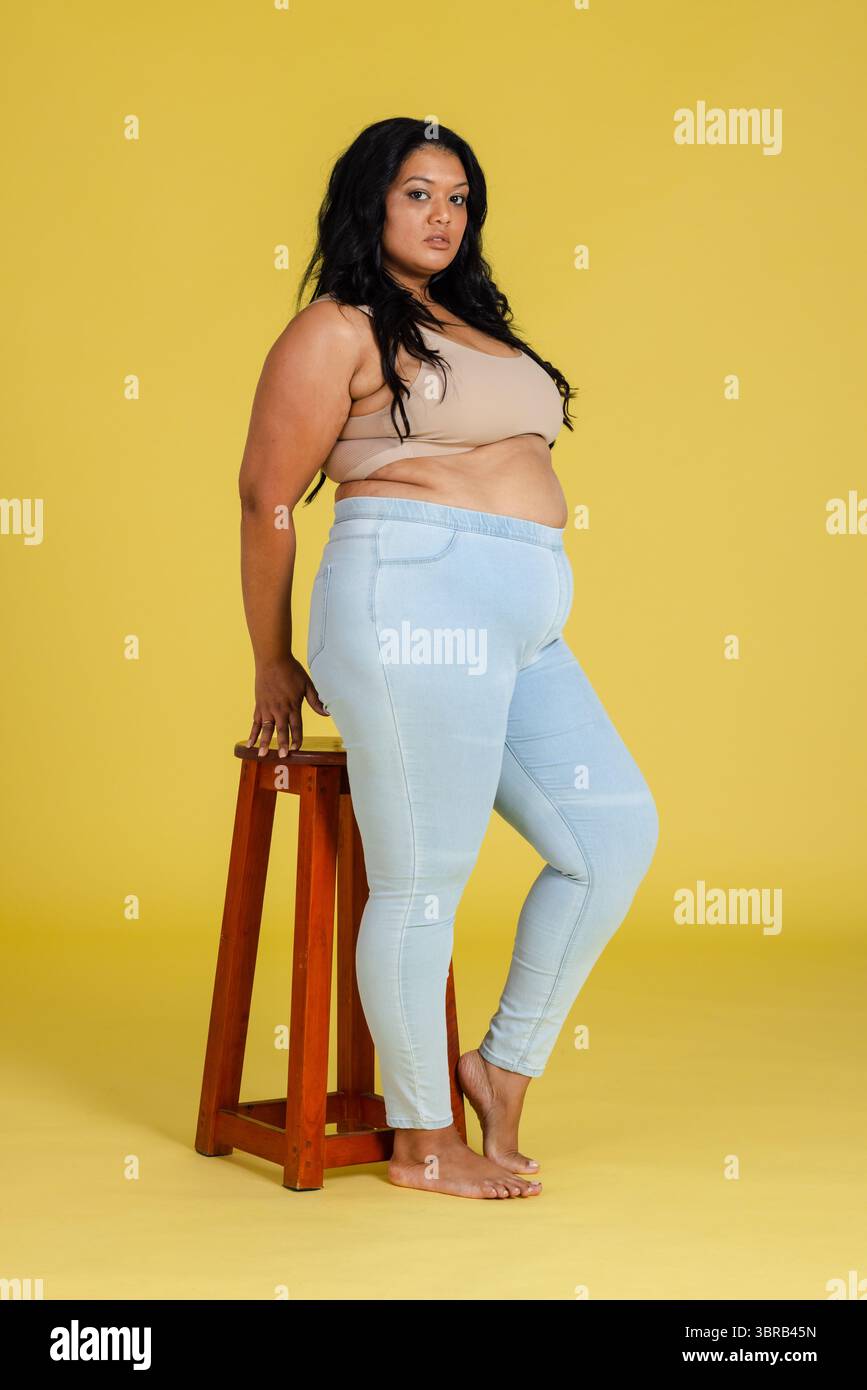 Femme posant en studio s'appuyant sur un tabouret en bois portant un crop top beige et un pantalon bleu Banque D'Images
