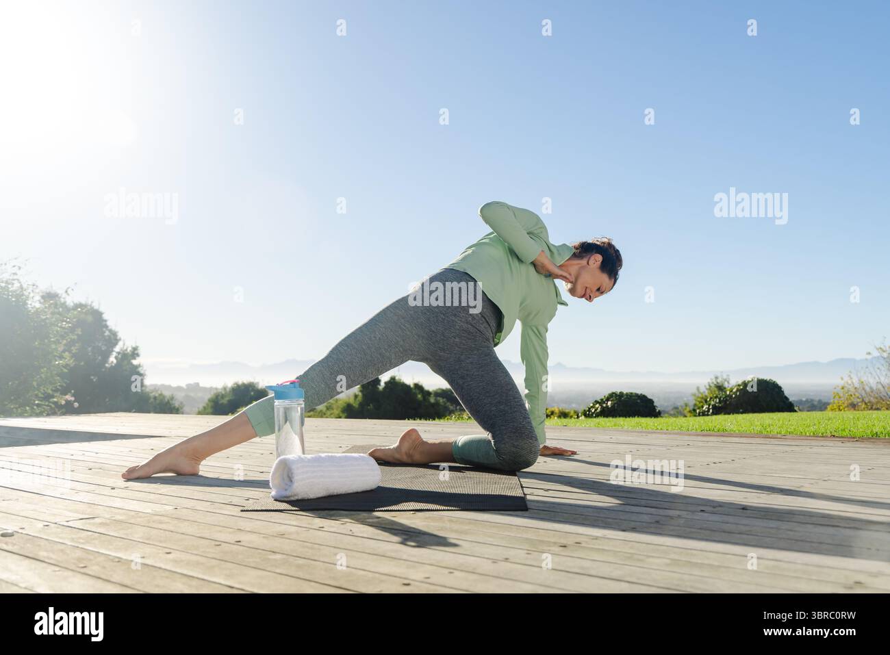 Femme adulte mature effectuant du yoga étirable latéral sur une terrasse en bois avec tapis, serviette et bouteille d'eau Banque D'Images
