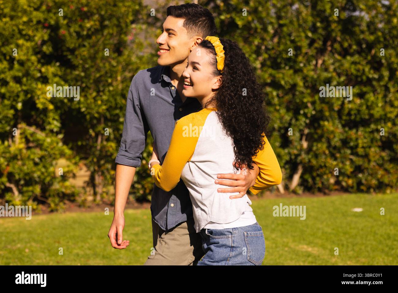 Couple diversifié embrassant et souriant sur une pelouse herbeuse par haie verte portant un bandeau jaune Banque D'Images