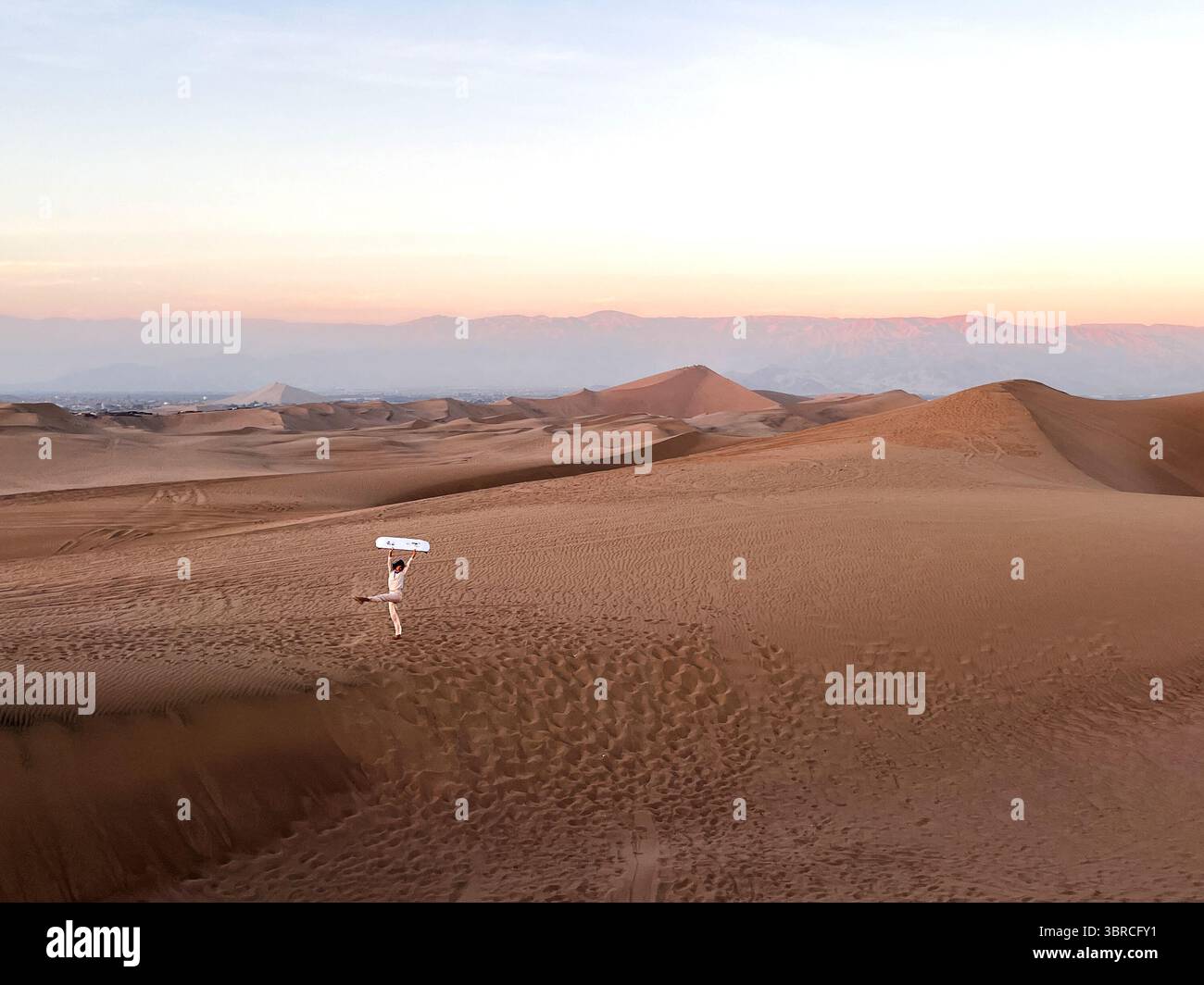 Femme en vêtements de couleur vive debout dans le désert sur les dunes et tenant la planche de sable au-dessus de la tête après avoir traîné sur le sable. Banque D'Images
