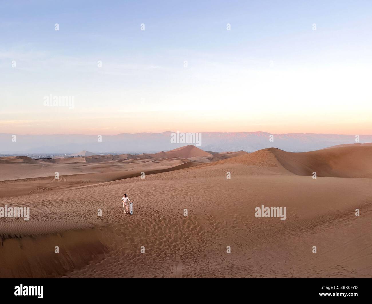 Femme en vêtements de couleur vive debout dans le désert sur les dunes et tenant la planche de sable à la main juste après avoir traîné sur le sable au Pérou, Huacachina. Banque D'Images