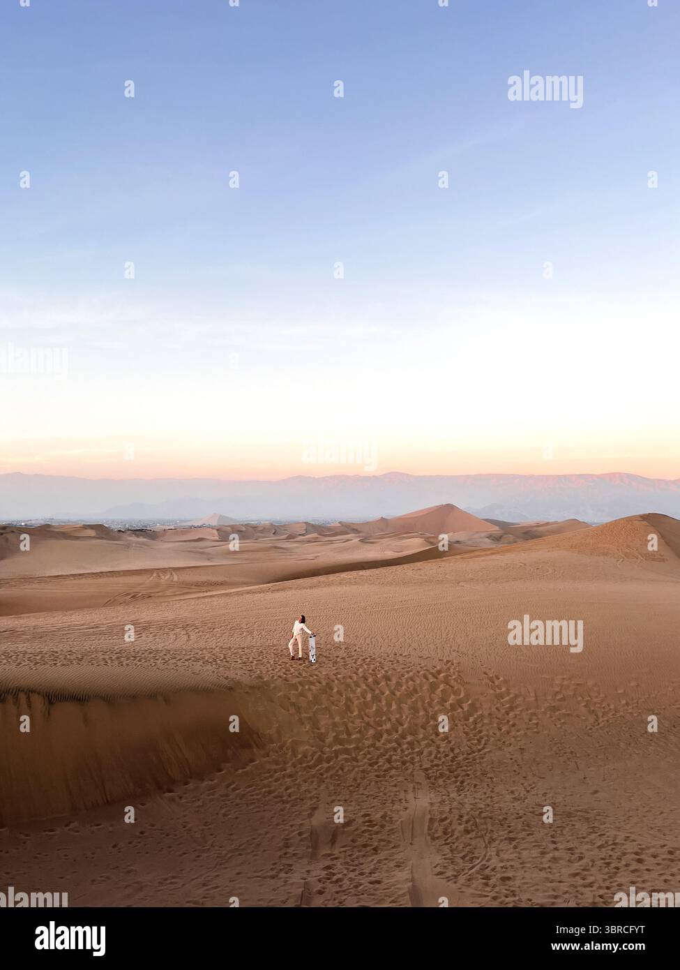 Photo verticale d'une femme en vêtements de couleur vive debout dans le désert sur les dunes et tenant la planche de sable à la main juste après avoir traîné sur le sable Banque D'Images