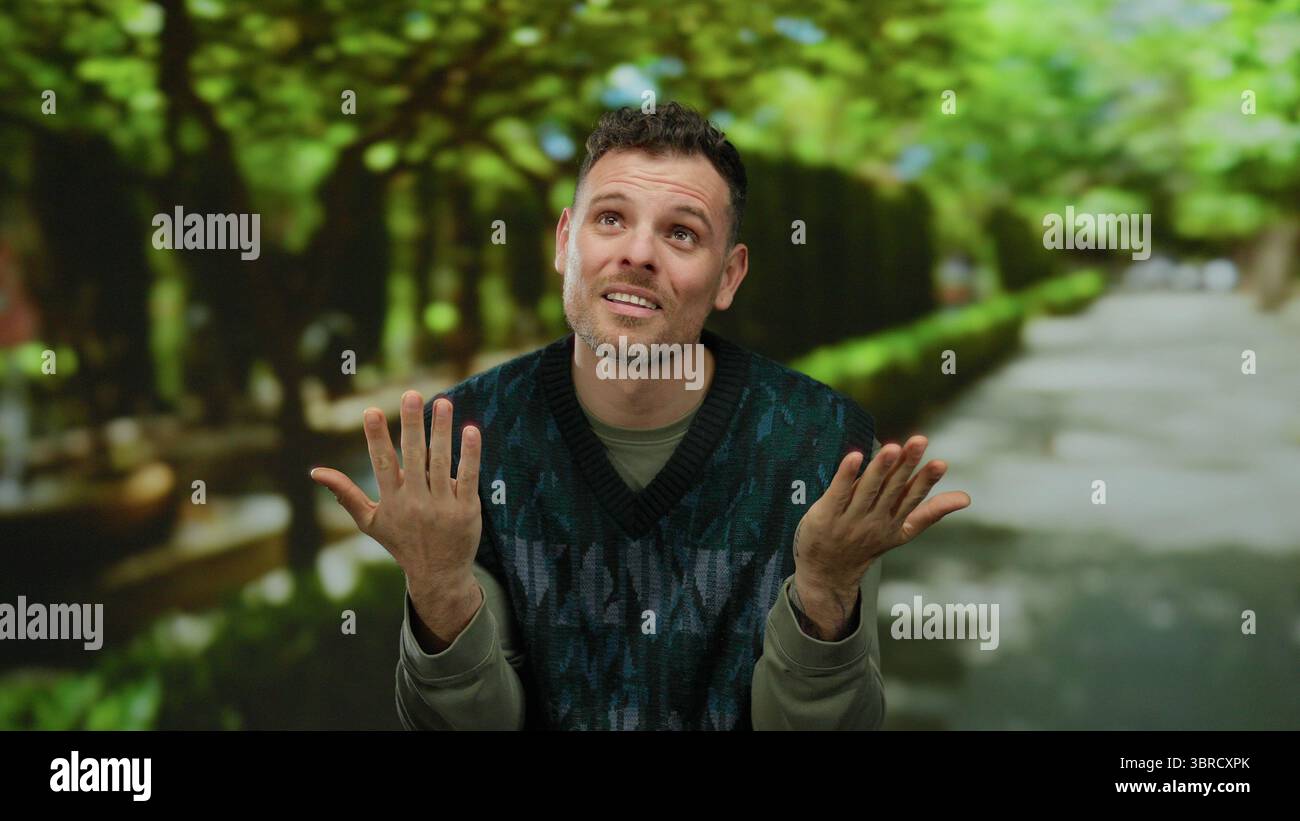 Homme haussant les épaules à l'extérieur vert parc tenue décontractée lumière du jour arbres flou arrière-plan détendu ambiance réfléchie portrait franc geste émotion naturel envi Banque D'Images