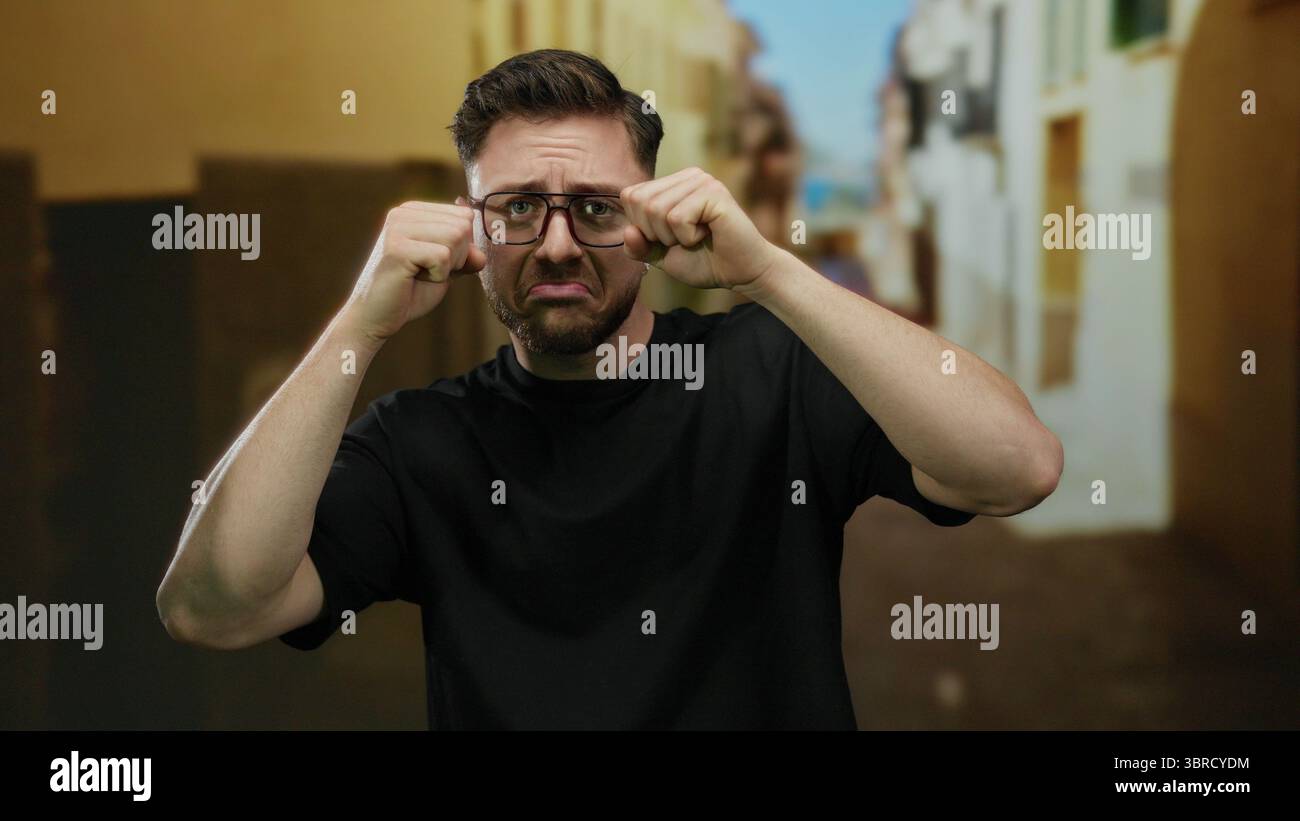 Jeune homme avec la barbe et les lunettes prétendant pleurer dans un cadre extérieur de rue de ville mettant en valeur l'expression ludique et l'émotion comique. Banque D'Images