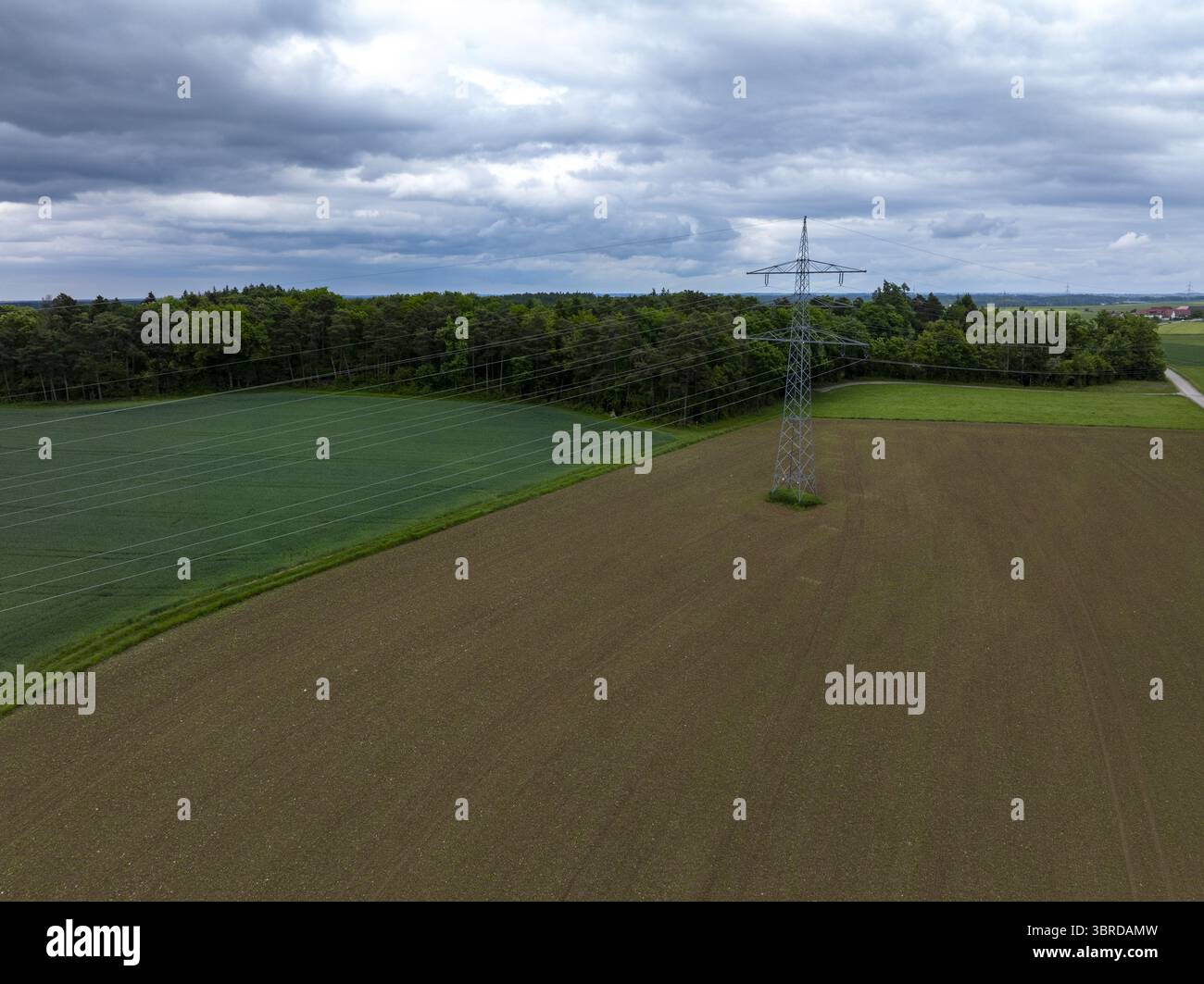 Vue aérienne d'un pylône électrique austère au milieu des textures contrastées de champs fraîchement labourés et de forêt verdoyante sous un ciel couvant, donc Banque D'Images