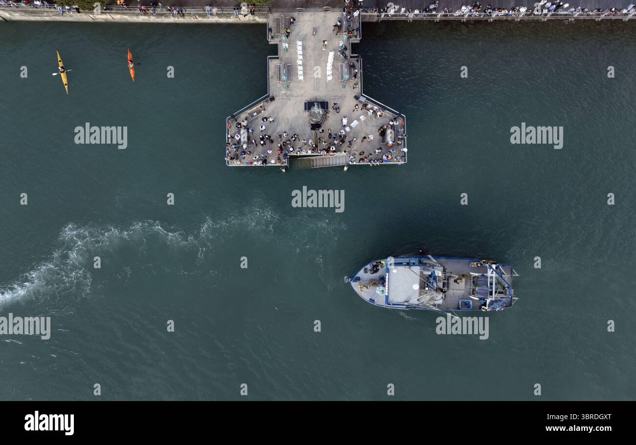 Vue aérienne d'une scène vibrante où un bateau coupe à travers les eaux sombres près d'un quai animé avec des gens, contrastant avec les kayaks à proximité, Nelson, région de Nelson, Nouvelle-Zélande. Banque D'Images