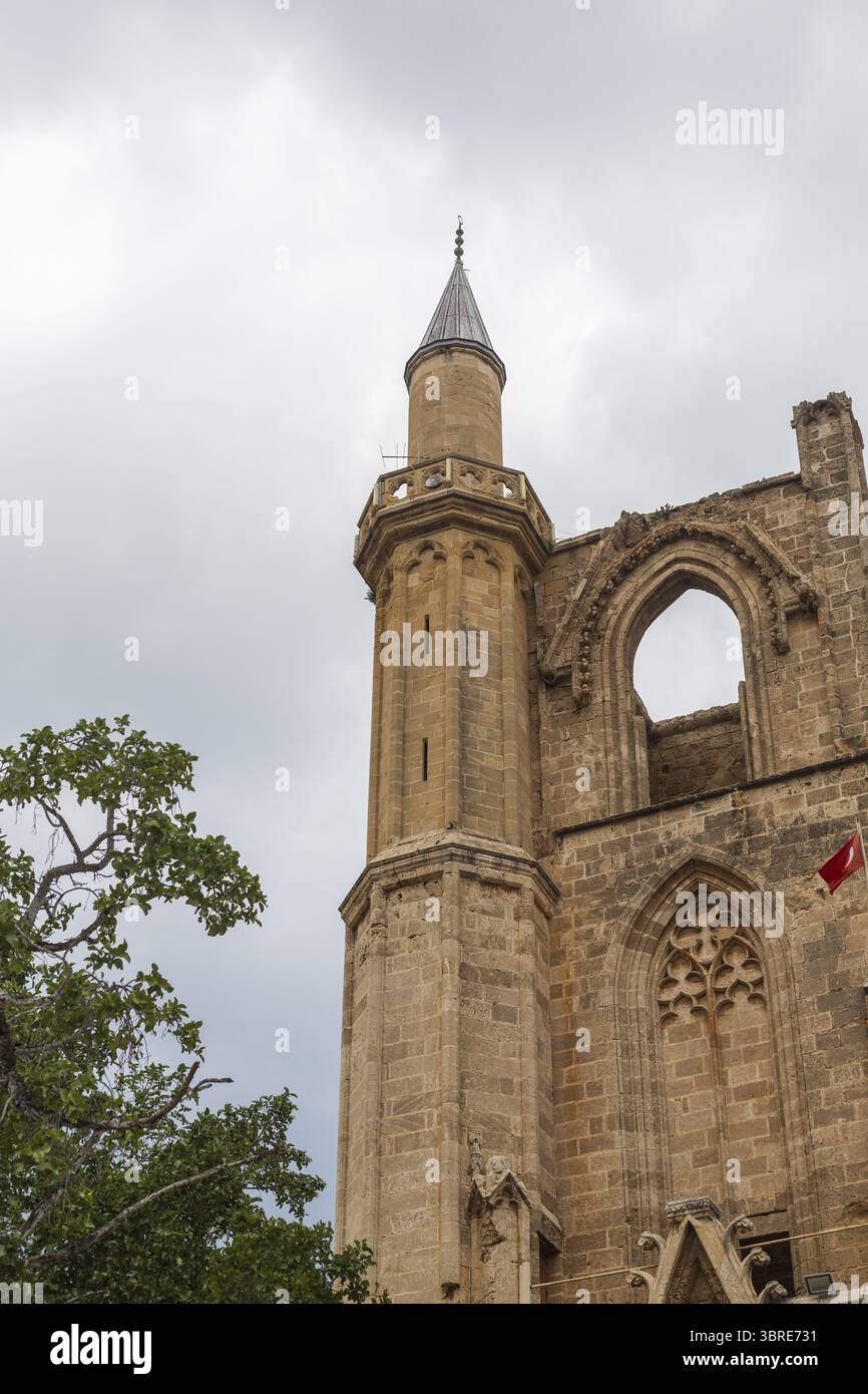 Vue d'un bâtiment en pierre altérée avec des fenêtres cintrées et une grande tour, encadrée par un ciel nuageux et des branches d'arbres, Varosha, Famagouste, Chypre. Banque D'Images