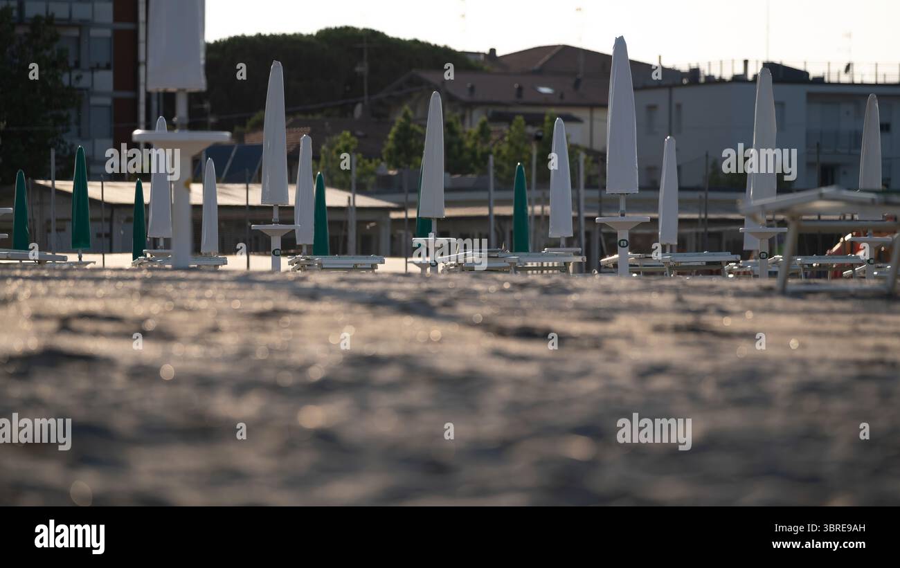 Des parasols fermés et des chaises longues vides se dressent en rangées sur une surface sablonneuse texturée près des bâtiments. Capture l'atmosphère calme avant les heures d'ouverture Banque D'Images