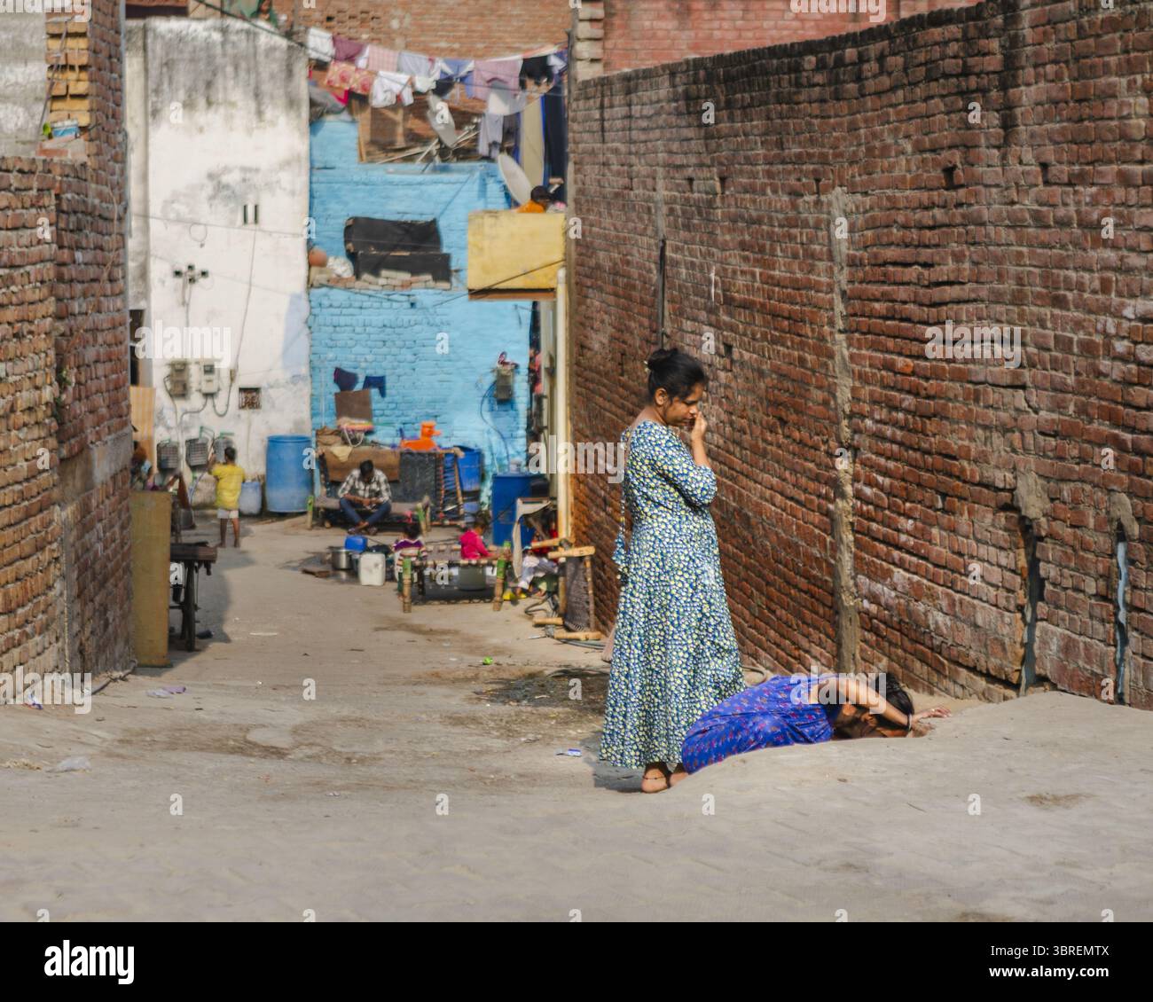 Agra, Inde - 24 juin 2025 : vue d'une femme dans une robe fluide debout près d'un mur de briques, observant une autre femme allongée sur le sol dans une ruelle étroite. Banque D'Images