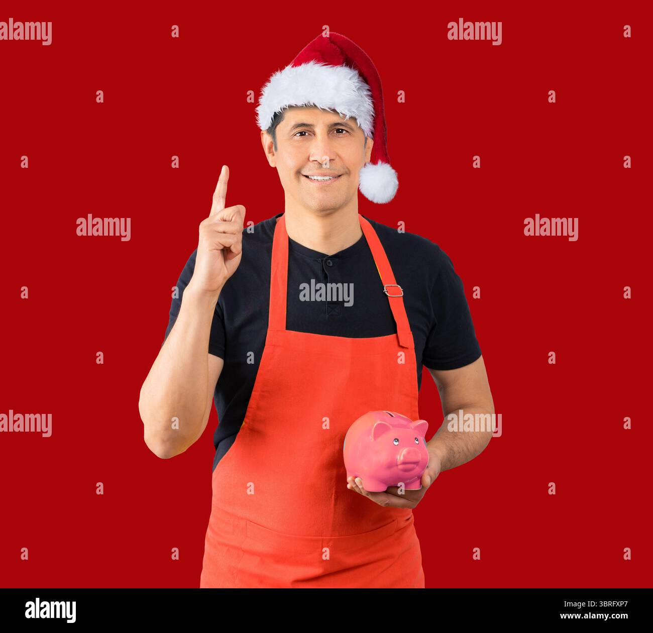 Photo de studio d'un homme portant un tablier rouge employé tenant une tirelire sur fond rouge tout en portant un chapeau de Noël. Banque D'Images