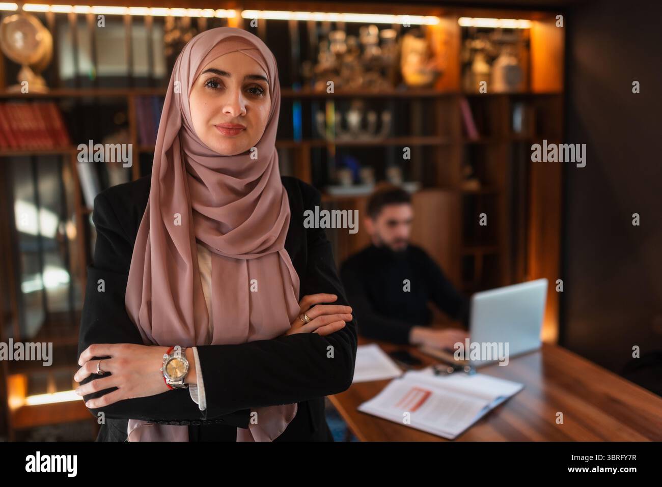 Femme d'affaires confiante portant un hijab debout avec les bras croisés dans un bureau moderne, un collègue masculin travaille sur un ordinateur portable en arrière-plan Banque D'Images