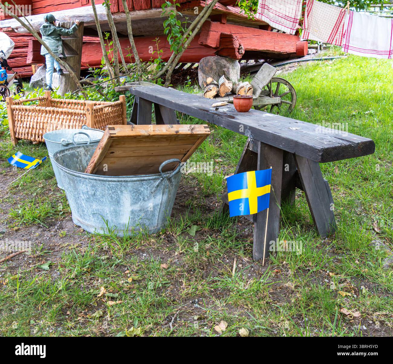Sodertalje, Suède - 6 juin 2025 : une scène extérieure rustique avec un banc en bois, des seaux en métal et des drapeaux suédois. Le cadre comprend de la verdure et Banque D'Images
