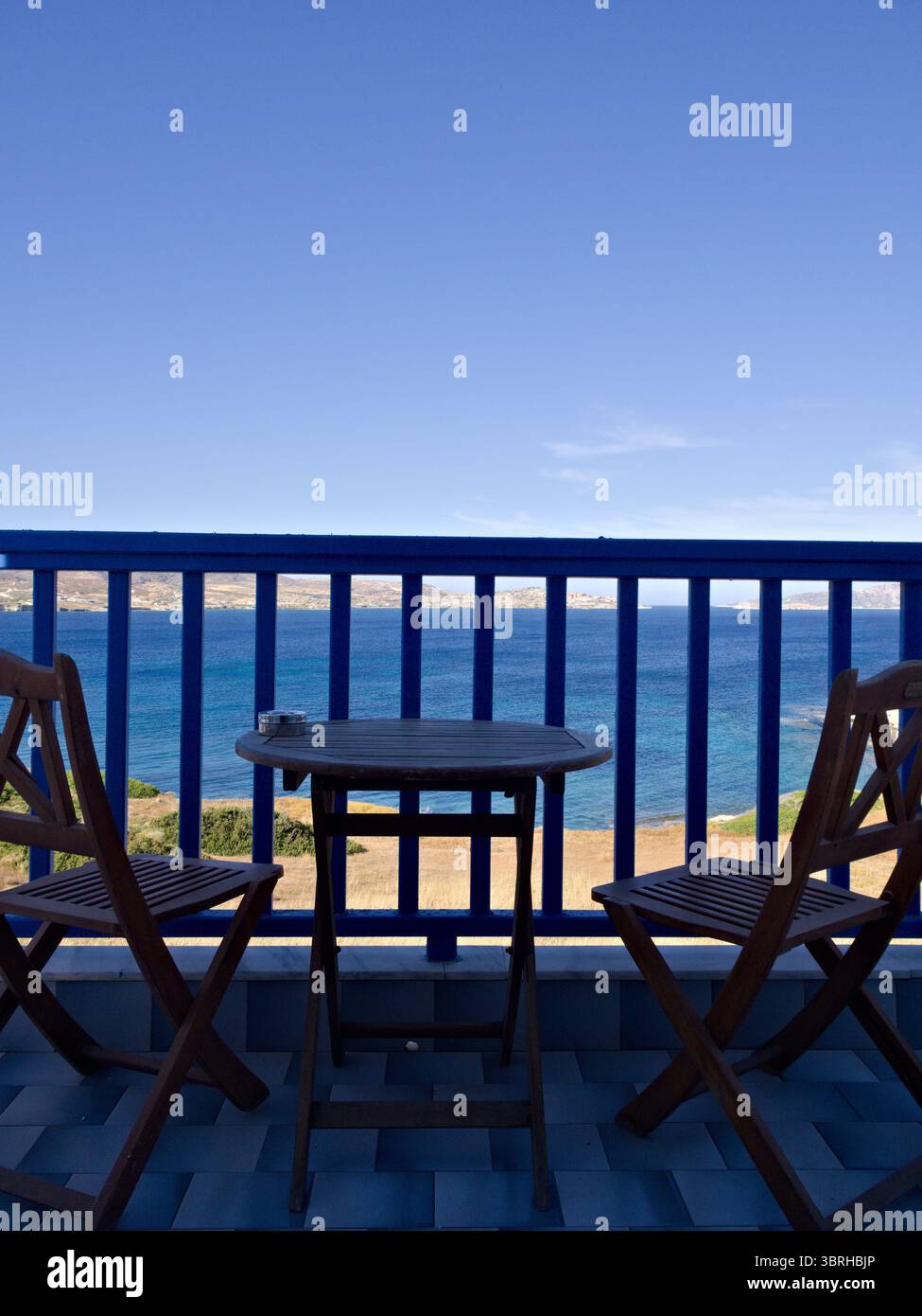 Balcon tranquille avec deux chaises en bois et une table face à la mer sous un ciel bleu vibrant. Idéal pour les voyages et les thèmes de style de vie. Banque D'Images