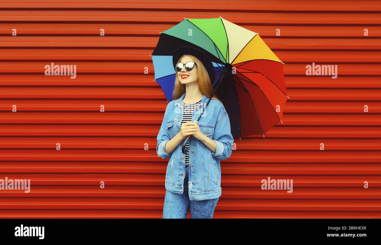 Portrait d'automne de jeune femme élégante et souriante tenant un parapluie coloré portant une veste de jean sur fond rouge Banque D'Images