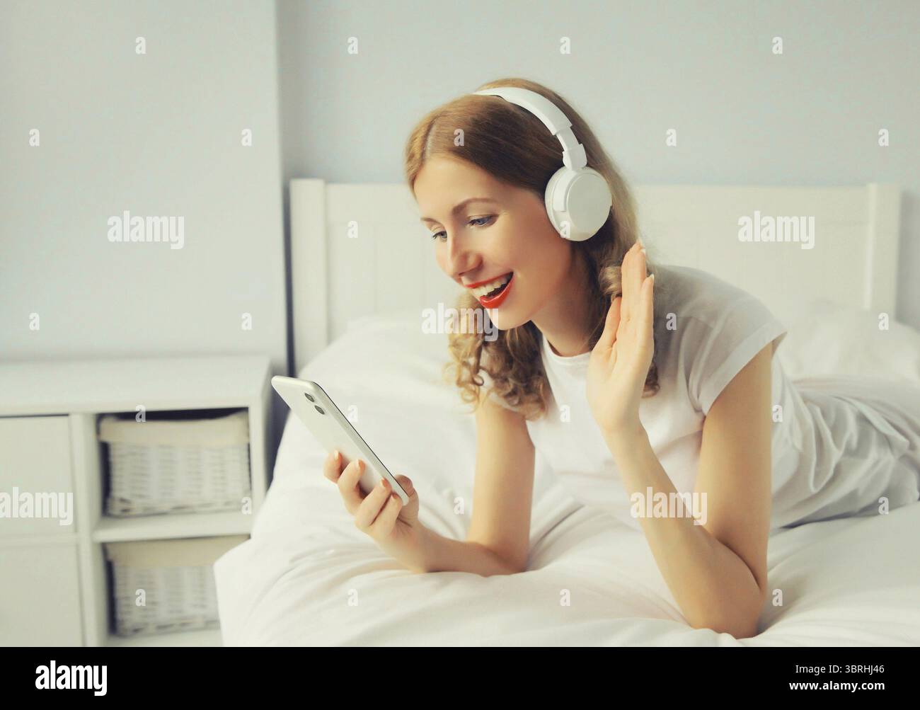Heureuse jeune femme souriante écoutant de la musique avec des écouteurs sans fil regardant smartphone parler sur appel vidéo sur le lit dans une chambre blanche à la maison Banque D'Images