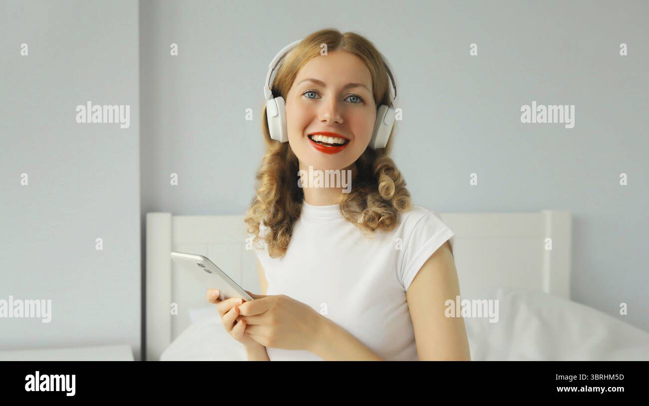 Heureuse jeune femme souriante écoutant de la musique avec des écouteurs sans fil regardant smartphone sur le lit dans une chambre blanche à la maison Banque D'Images