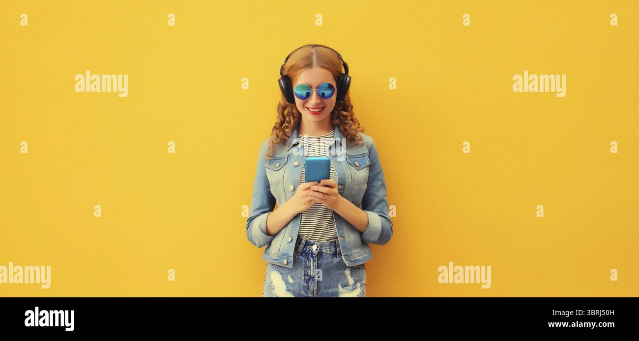 Portrait de jeune femme heureuse moderne élégante écoutant de la musique dans des écouteurs avec une veste de port de téléphone, lunettes de soleil sur fond jaune Banque D'Images