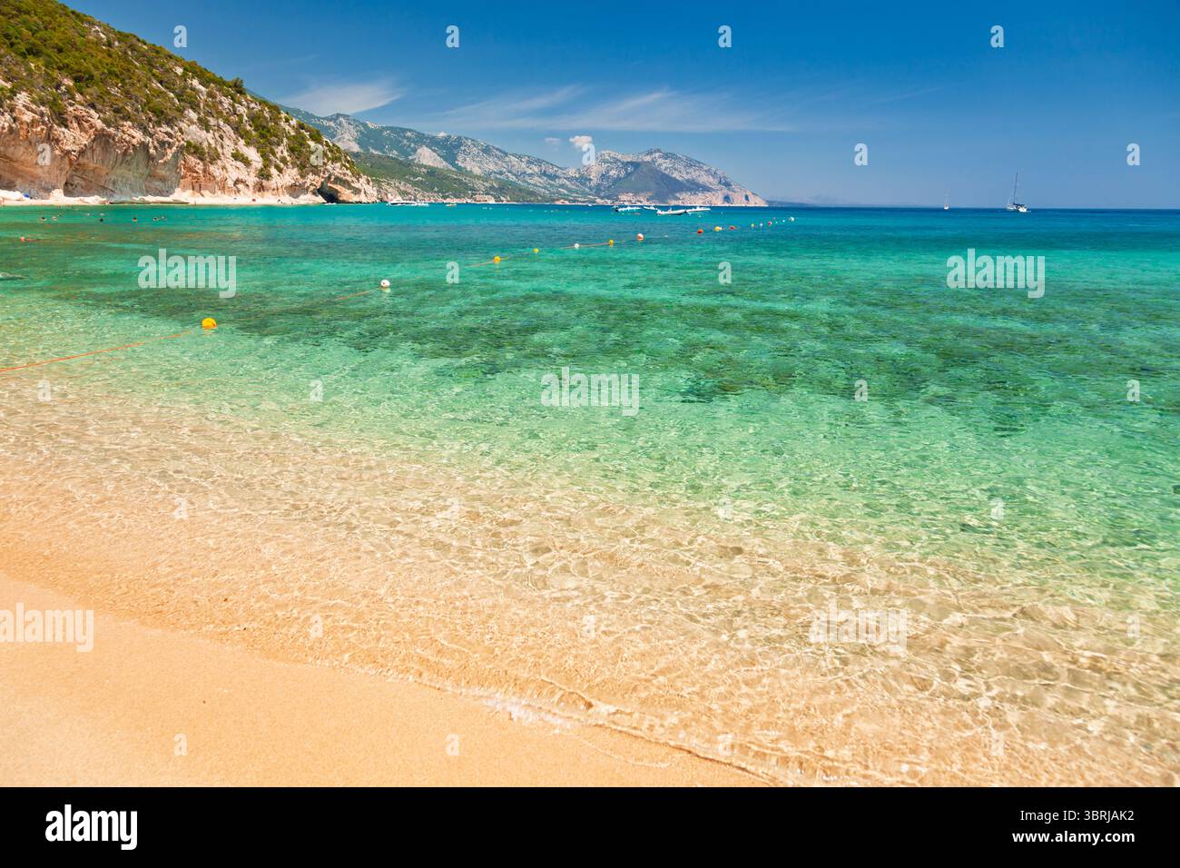 La plage de Cala Luna, sur la côte est sarde, présente une eau turquoise peu profonde et limpide qui rencontre doucement du sable doré, avec des bateaux ancrés dans la distance Banque D'Images