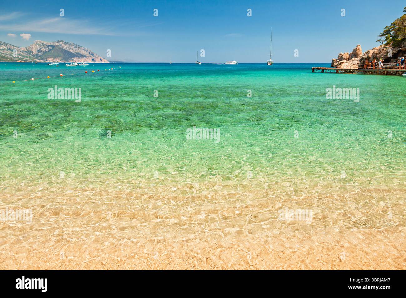 La plage de Cala Luna, sur la côte est sarde, présente une eau turquoise peu profonde et limpide qui rencontre doucement du sable doré, avec des bateaux ancrés dans la distance Banque D'Images