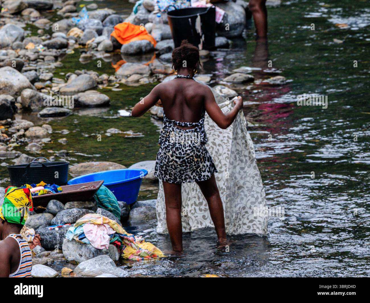 villageois local en robe à imprimé léopard est debout au fond de la cheville dans une rivière peu profonde lavant un grand drap Banque D'Images