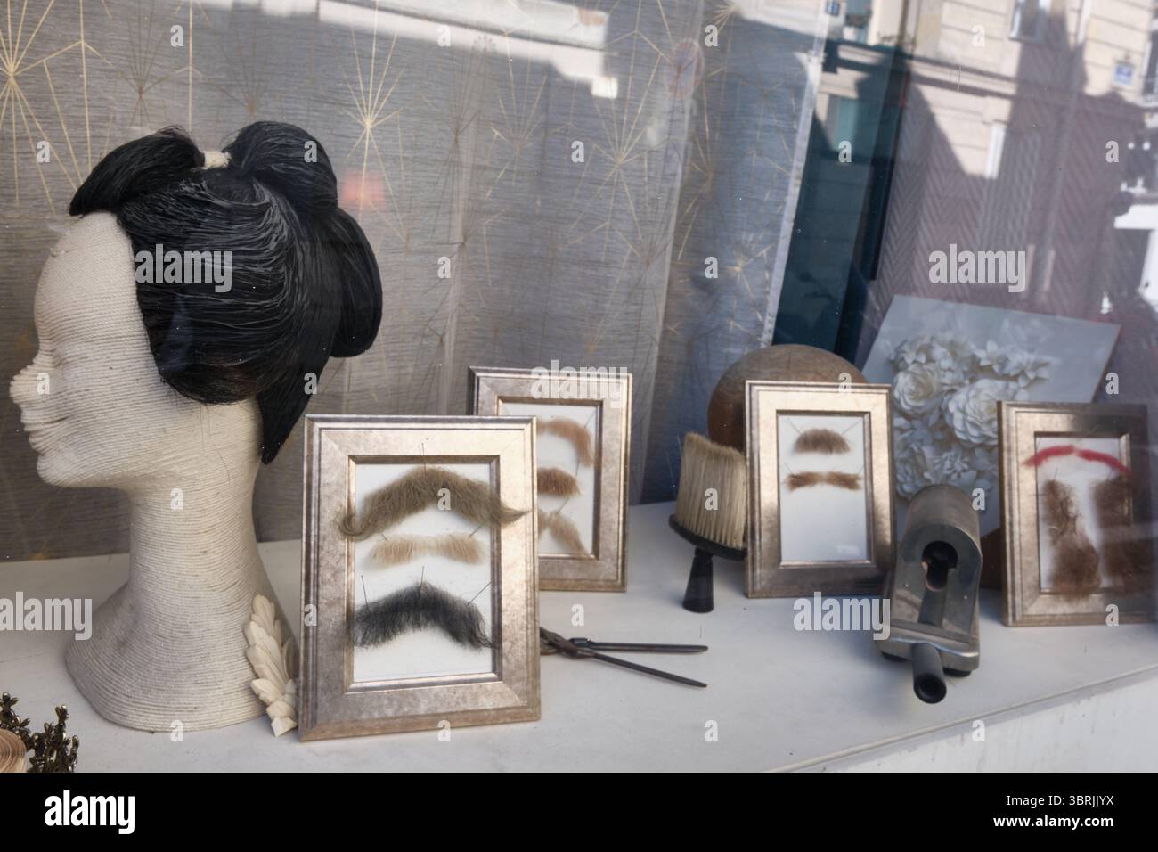 Vitrine présentant une tête de mannequin avec perruque entourée d'échantillons de moustache encadrés et d'outils de barbier au 10 Rue Rampon, Paris, France Banque D'Images