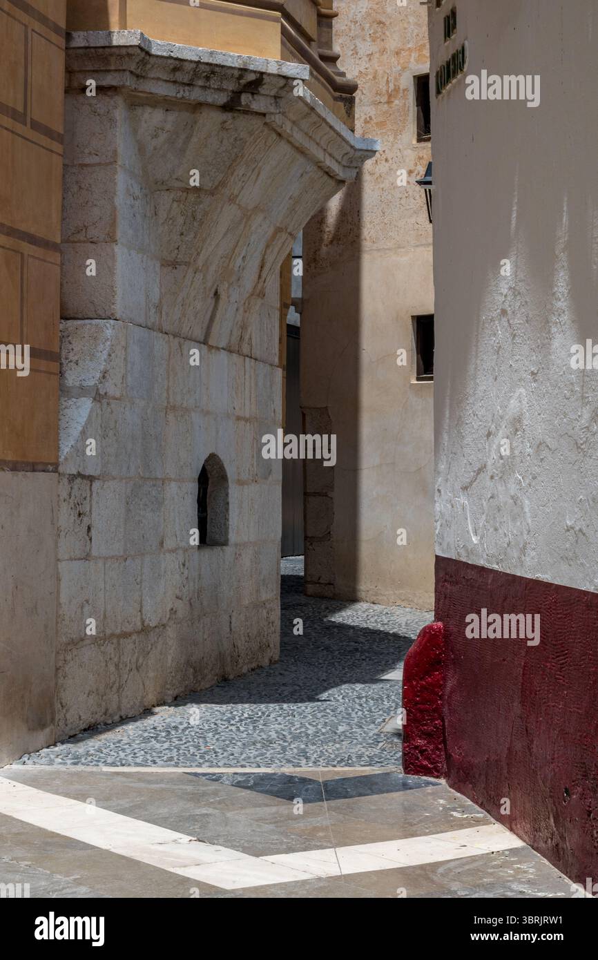 Une ruelle étroite entre des bâtiments en pierre baignés de soleil, révélant les textures anciennes de l’architecture historique de Málaga. Banque D'Images