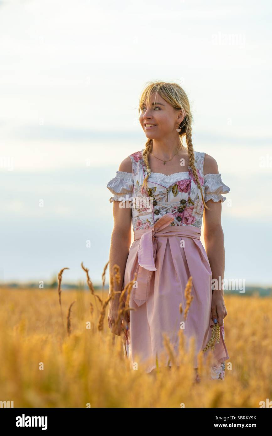 Charmante jeune femme portant une robe traditionnelle bavaroise dirndl avec des cheveux tressés, debout dans un champ de blé doré et regardant au loin. Vie rurale Banque D'Images